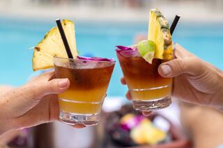 Two tropical cocktails clink together, garnished with pineapple and colorful flowers, set against a vibrant pool backdrop.