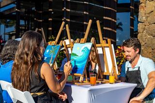A group of people enjoy a painting session outdoors, creating beach-themed artwork on easels, with drinks on the table.