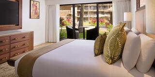 A cozy hotel room features a plush bed, decorative pillows, and large windows showcasing a vibrant outdoor view with lush greenery.