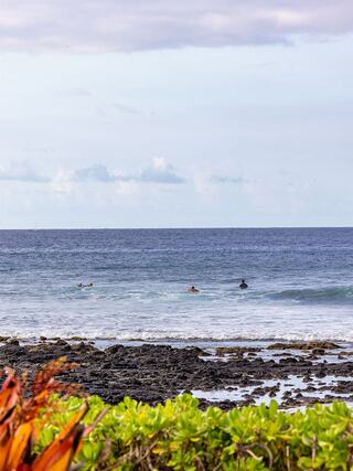 Surfers enjoy the waves off a rocky coastline, framed by vibrant greenery under a cloudy sky.