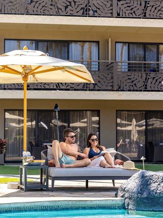 A couple lounges poolside under a large umbrella, enjoying each other's company with a modern hotel backdrop. Relaxation is the vibe.