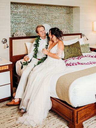 A joyful couple dressed in wedding attire sits on a bed, adorned with flowers and enjoying each other's company, surrounded by elegance.