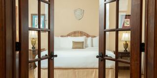 A cozy bedroom with a large bed, decorative headboard, warm lamps, and soft colors, inviting relaxation through double doors.