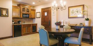 A cozy dining area features a round table with blue chairs, a wooden cabinet, a small kitchen space, and elegant decor on the walls.