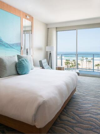 A bright, modern hotel room features a large bed and ocean views through expansive windows, creating a serene beachside atmosphere.