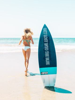 A woman in a bikini runs toward the ocean, carrying a surfboard with a motivational message, surrounded by a sunny beach backdrop.