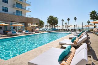 A clear blue pool is surrounded by lounge chairs and palm trees, with a sunny sky and a beachfront view in the background.