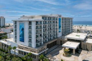 A contemporary beachfront hotel features modern architecture, a large mural of ocean waves, and a welcoming entrance amidst lush greenery.