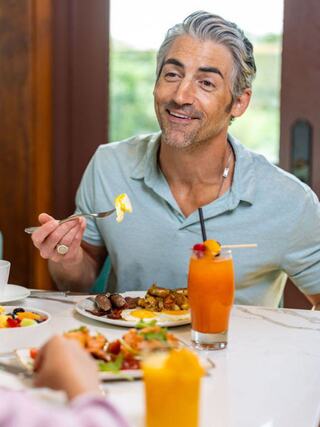 A man enjoys a cheerful breakfast, smiling while holding a fork with food. Colorful drinks and a variety of dishes are on the table.