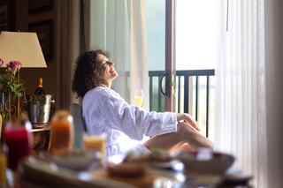 A woman in a white bathrobe enjoys a moment of relaxation by the window, sipping a drink with a spread of breakfast items nearby.