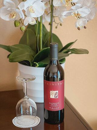 A bottle of Trinitas wine sits next to a wine glass, surrounded by elegant white orchids in a stylish pot on a polished table.