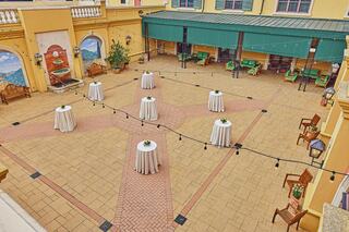 An outdoor courtyard features several round tables with light linens, surrounded by chairs and decorative elements, under string lights.