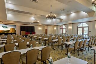 A spacious conference room featuring rows of tables and chairs, with a large presentation screen and windows letting in natural light.