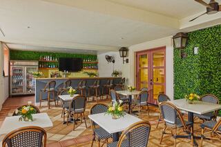 A cozy dining area features wicker chairs and tables with floral centerpieces, a bar with drinks, and greenery decor across the walls.