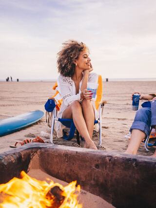A woman with curly hair sits by a beach bonfire, enjoying a drink and smiling, with a surfboard nearby and friends chatting.