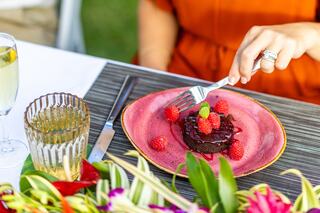 A dessert plate features a chocolate treat garnished with fresh raspberries and mint, accompanied by drinks and colorful floral decorations.