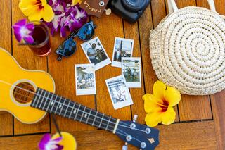 A vibrant tropical scene with a ukulele, sunglasses, a woven bag, polaroid photos, a drink, and colorful flowers, evoking a joyful summer vibe.