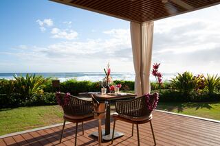 A cozy dining setup on a patio with ocean views, adorned with tropical flowers, surrounded by lush greenery and a bright, sunny sky.
