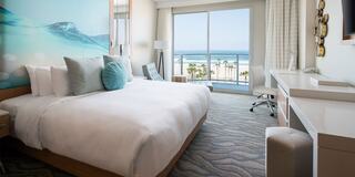 A stylish hotel room features a large bed, a turquoise accent pillow, modern furnishings, and a stunning ocean view through large windows.