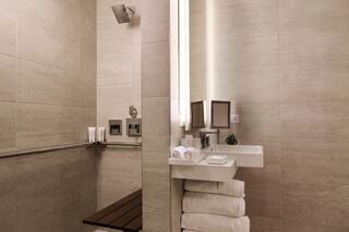 A modern bathroom features neutral tiles, a rainfall shower, wooden bench, and neatly arranged towels, exuding a clean, serene ambiance.