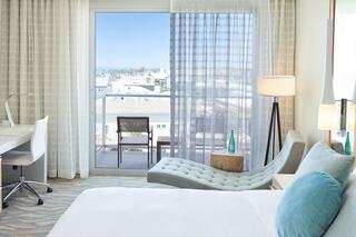 A modern hotel room features a cozy bed, stylish furniture, and large windows with a view, bathed in natural light.