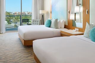 Bright, modern hotel room with two beds, colorful pillows, a cozy chair, and large windows showcasing palm trees outside.