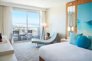 A modern hotel room features a comfortable bed, a chaise lounge, and a stunning ocean view from the balcony. Elegant decor and light colors create a serene atmosphere.