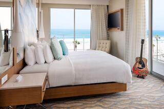 A modern coastal hotel room features a plush bed, large windows with ocean views, and a guitar resting against a chair.