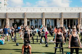A large group participates in an outdoor fitness class, using weights on a grassy area, against a backdrop of modern architecture and clear skies.
