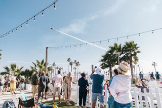 A crowd gathers outdoors, watching a jet perform acrobatics in the sky, surrounded by palm trees and festive decorations.