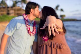 A couple kisses on a beach, with the woman displaying her engagement ring, surrounded by a tropical setting.