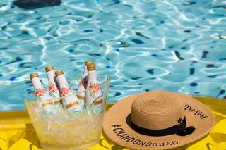 Chilled bottles of sparkling wine in an ice bucket next to a stylish straw hat, all resting by a vibrant blue pool.