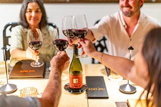 Four people clink glasses filled with red wine at a table, celebrating together. A bottle of wine sits prominently in the center.