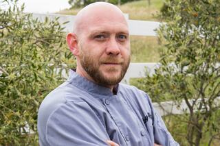 A bald man with a beard stands confidently in a chef's coat, posed against a backdrop of greenery and a white fence.