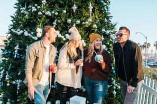 Two people in festive sweaters stroll past a large Christmas tree during an outdoor ice skating event, surrounded by cheerful holiday lights.