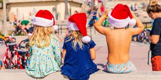 Three children wear Santa hats while sitting on a ledge, enjoying a festive atmosphere. Bicycles and people are nearby in a lively setting.