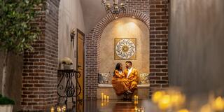 A couple in matching traditional outfits sits together in a cozy, softly lit corridor with candles, creating an intimate atmosphere.
