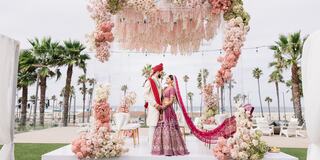 A couple in traditional attire stands under a floral arch, surrounded by palm trees and ocean views, exuding romance and celebration.