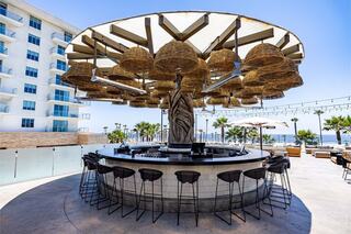 A stylish outdoor bar features a circular design with hanging woven shades, surrounded by modern seating and palm trees in a sunny setting.