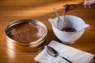 A round tin of baked chocolate dessert sits next to a bowl of chocolate mix and whisk, on a wooden surface with a spoon and napkin.