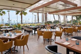 A stylish restaurant features elegant seating, palm trees, and a scenic view of the ocean, creating a relaxed dining atmosphere.