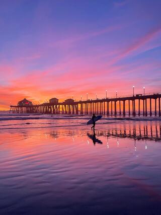 A couple strolls hand-in-hand along the beach, smiling and enjoying the sunny day with blue skies and gentle waves in the background.