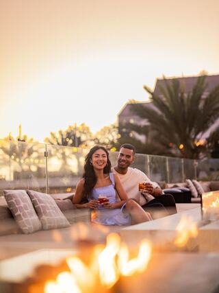 A happy couple enjoys drinks together on a cozy outdoor couch, with a warm sunset and fire enhancing the romantic atmosphere.