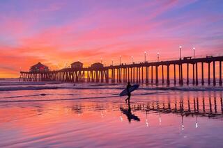 A surfer walks along the shore at sunset, with vibrant pink and orange hues reflecting off the water near a lit pier.