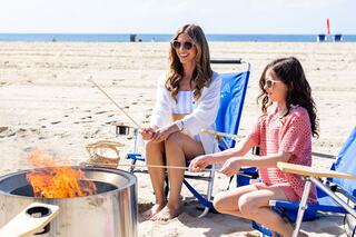 Two women sit near a beach fire, roasting marshmallows. They enjoy a sunny day, wearing sunglasses, with beach chairs and a basket nearby.