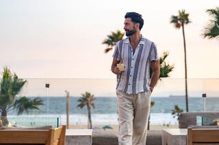 A man in a stylish shirt holds a drink while enjoying a sunset view of the ocean and palm trees from a modern terrace.