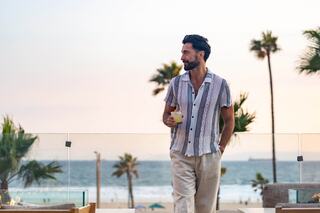 A man in a stylish shirt walks along a beachside terrace, holding a drink, with palm trees and the ocean in the background at sunset.