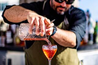 A bartender pours a vibrant red cocktail into a stylish glass, showcasing a sunny outdoor setting with various bottles in the background.