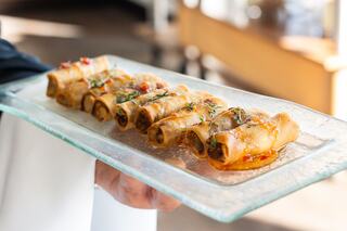 A glass platter holds neatly arranged, golden-brown rolled tacos topped with sauce and herbs, ready to be served.