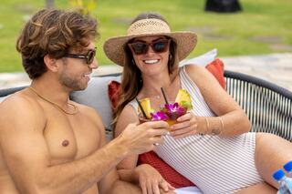 A cheerful couple enjoys tropical drinks by the pool, surrounded by lush greenery, embracing a relaxed, sunny day together.